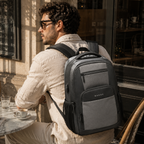 A man wearing a gray and black RANGE backpack sits at a cafe table, viewed from behind as he looks out the window.