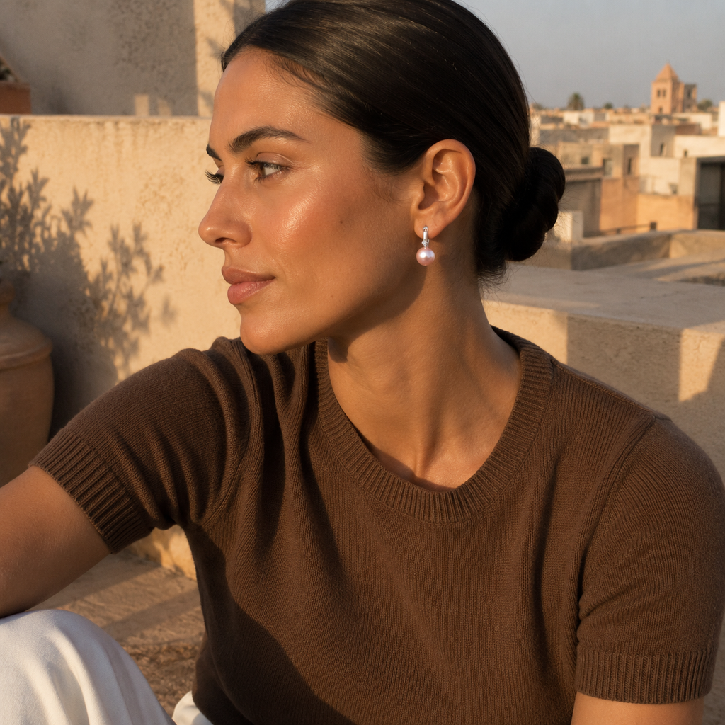 A woman in a brown knit short-sleeve top wearing a single dangling pearl earring, photographed outdoors on a sunlit rooftop with buildings in the background.
