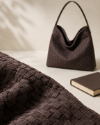 A dark brown woven leather shoulder bag with a single top handle, styled on a light beige surface next to a book, with a matching woven fabric prop in the foreground.