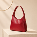 A red leather shoulder bag with a single top handle and a front slip pocket, displayed on a light beige pedestal against a warm beige background with a subtle gold circular wall decor.