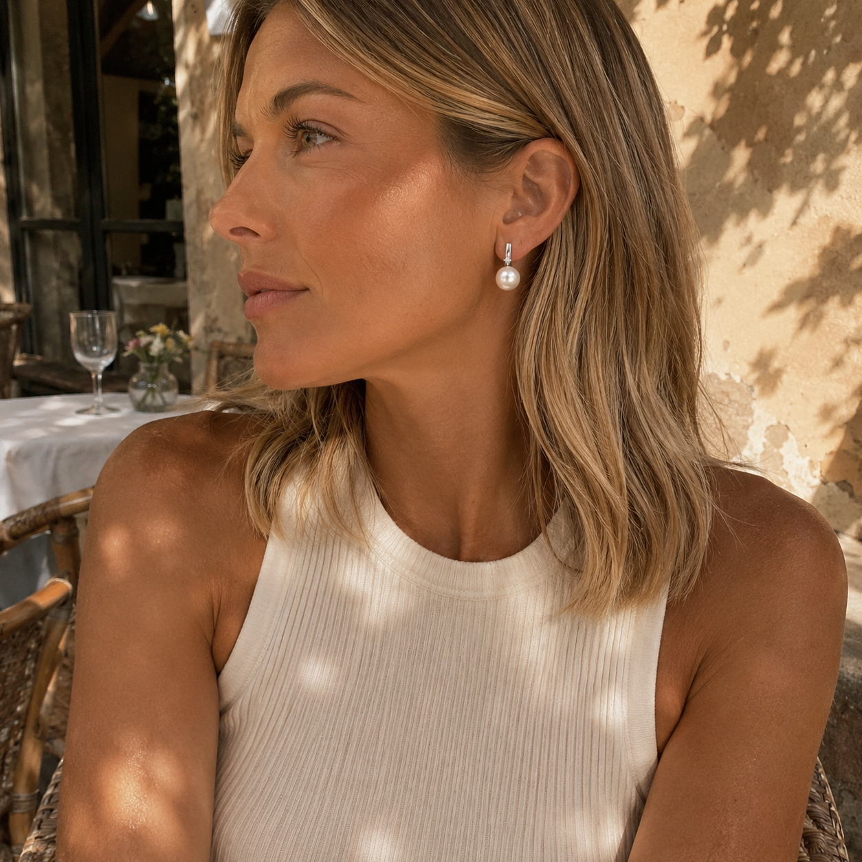 A woman with blonde hair wearing a white ribbed sleeveless top and a single white pearl drop earring, photographed in a sunlit outdoor setting.