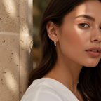 A close-up lifestyle shot of a woman wearing a delicate gold huggie hoop earring with small clear stones, set against a warm stone pillar background.