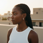 A woman with braided hair wearing a white ribbed tank top and a small silver hoop earring, photographed outdoors against a softly blurred beige building background.