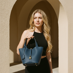 A blonde woman in a black sleeveless top holds a structured navy blue leather handbag with gold-tone hardware and a front lock detail, standing in front of a beige arched architectural background.