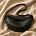 A black pebbled leather crescent-shaped shoulder bag with a curved flap and a thin strap, styled on a draped beige fabric background.