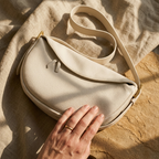 A cream-colored pebbled leather crescent-shaped shoulder bag with a matching strap, styled on a textured beige linen cloth with warm sunlight and shadows. A hand with a gold ring rests on the bag.