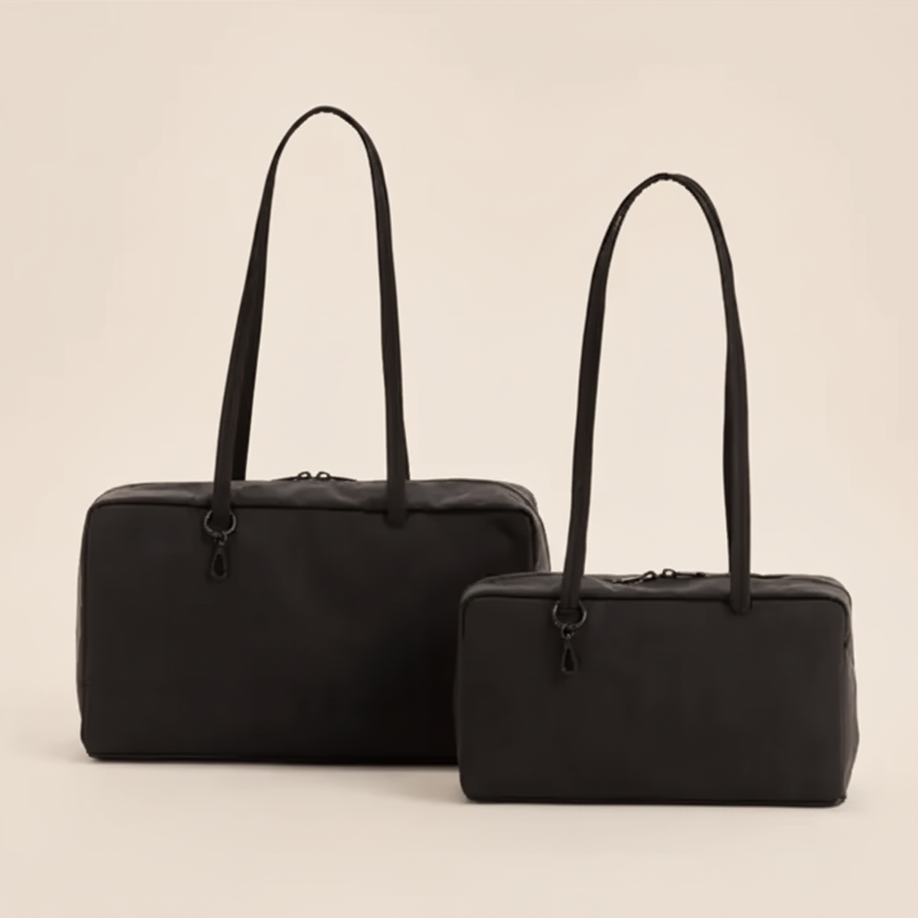 Two black leather duffel bags with long shoulder straps, one large and one small, displayed on a warm beige background.