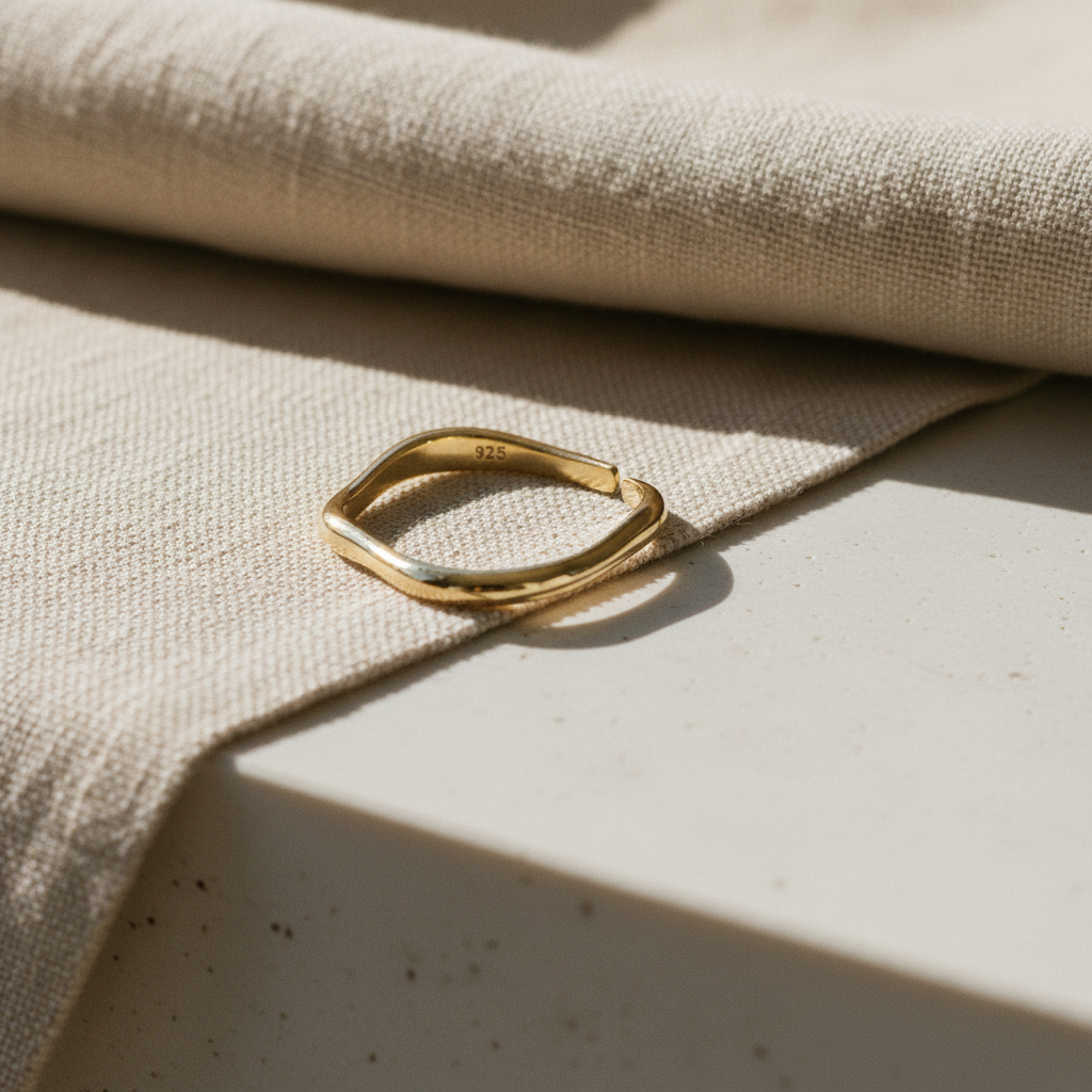 A polished gold ring with a wavy, organic open shape resting on a textured beige linen cloth over a light stone surface.