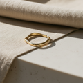 A polished gold ring with a wavy, organic open shape resting on a textured beige linen cloth over a light stone surface.