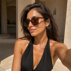 A woman wearing tortoiseshell sunglasses and a black halter top, taking a selfie in a sunlit architectural setting with beige stone walls.