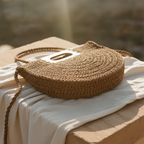 A round, handwoven straw bag with a braided handle, resting on draped cream-colored fabric atop a wooden surface in warm, sunlit outdoor light.