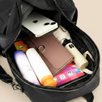 A black backpack shown open from above, filled with everyday items including a white smartphone, a brown wallet, a cream-colored umbrella, a purple and white bottle, and other accessories, set against a solid light beige background.