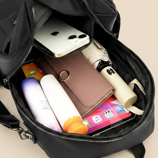 A black backpack shown open from above, filled with everyday items including a white smartphone, a brown wallet, a cream-colored umbrella, a purple and white bottle, and other accessories, set against a solid light beige background.