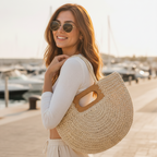 A woman wearing round sunglasses and a white ribbed long-sleeve top carries a large round woven straw tote bag with brown handle accents, set against a blurred marina background at sunset.