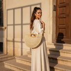 A woman in a long white dress stands on stone steps outside a building, carrying a large woven straw tote bag with wooden handles.