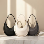 Three crescent-shaped shoulder handbags in black, off-white, and dark gray, arranged on a white marble surface with warm sunlight casting shadows against a beige wall.