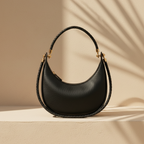 A black pebbled leather crescent-shaped shoulder bag with a braided top handle and gold-tone hardware, displayed on a beige platform with soft shadow overlays.