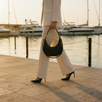 A person in a cream-colored pantsuit walks along a waterfront promenade at sunset, holding a black crescent-shaped handbag with gold-tone hardware. Boats and water are visible in the background.