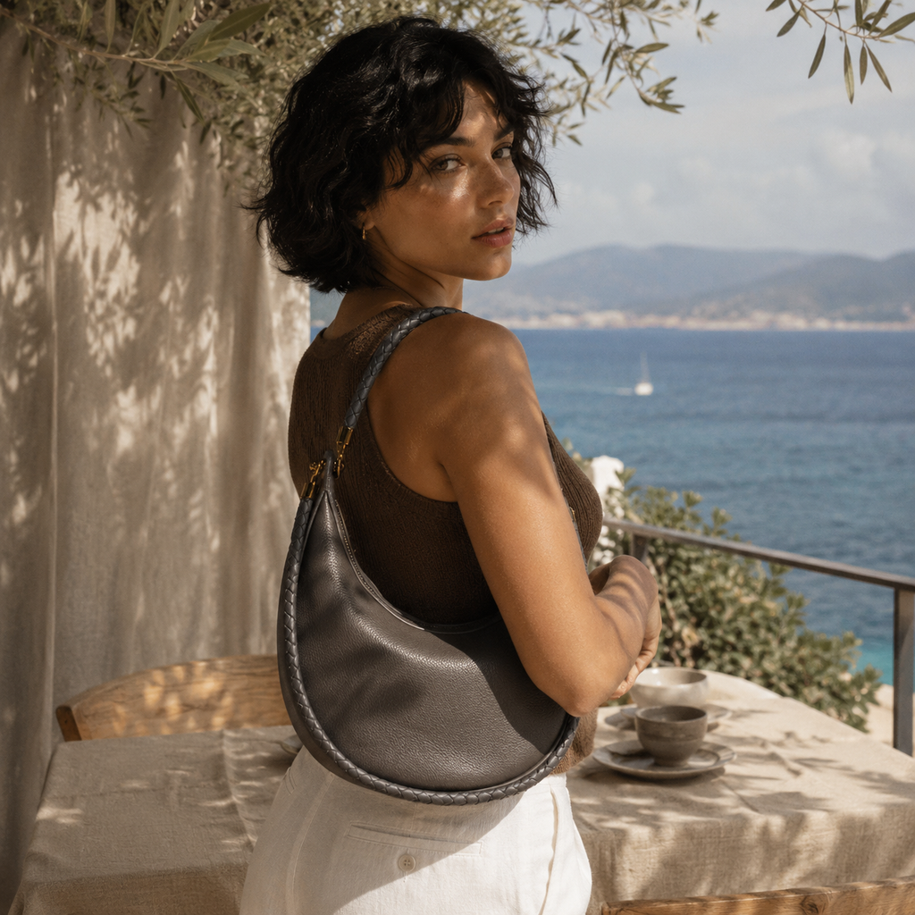 A woman with short dark hair stands on a sunlit balcony overlooking the blue sea, wearing a taupe leather crescent shoulder bag with braided trim. She is dressed in a brown top and white pants, with a table and cups beside her.