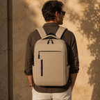 A man viewed from behind wearing a light beige backpack with black accents, standing against a textured beige wall with warm sunlight and leaf shadows.